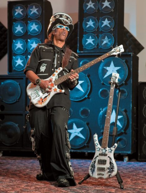 Bootsy Collins mit großem Hut und Sonnenbrille spielt einen Warwick Infinity-Bass. Daneben steht ein weiterer Bass auf dem Boden.